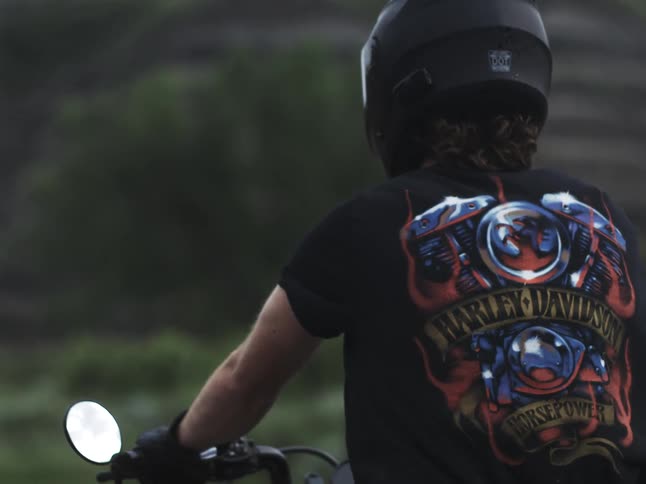 Man wearing a t shirt with a harley davidson design sits on a motorcycle on a summer day