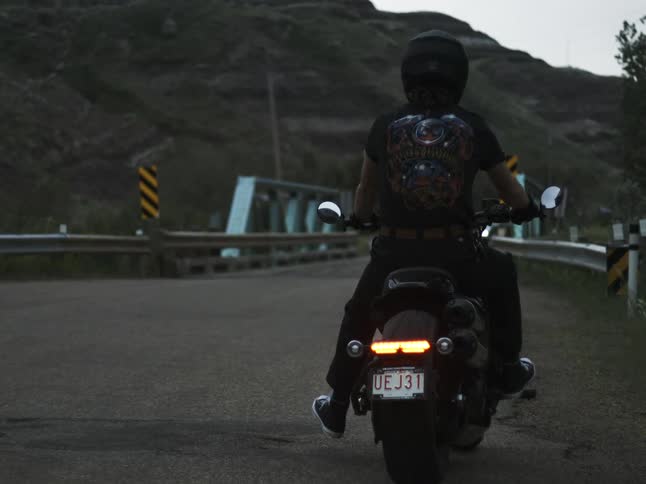 A biker with a helmet and a t shirt featuring a unique graphic sits on a motorcycle on a country road at dusk or dawn with a bridge and hills in the background