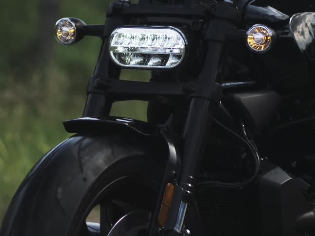 A close view shows the front wheel forks and handlebars of a black motorcycle with its bright led headlight and blinking orange turn signals illuminated on a roadside in daylight