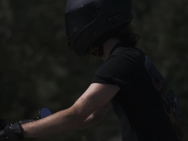 A person wearing a black helmet and dark tshirt rides a motorcycle visible from the shoulder to the forearm against a dark blurred background on a sunny day