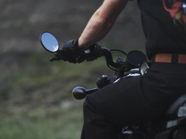 A gloved rider in a graphic tee grips a black motorcycles bars round mirrors and gauges set against a blurred green backdrop