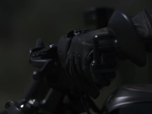 A person wearing a black motorcycle glove grips the throttle and handlebar of a black motorcycle in a close view with a dark blurred background