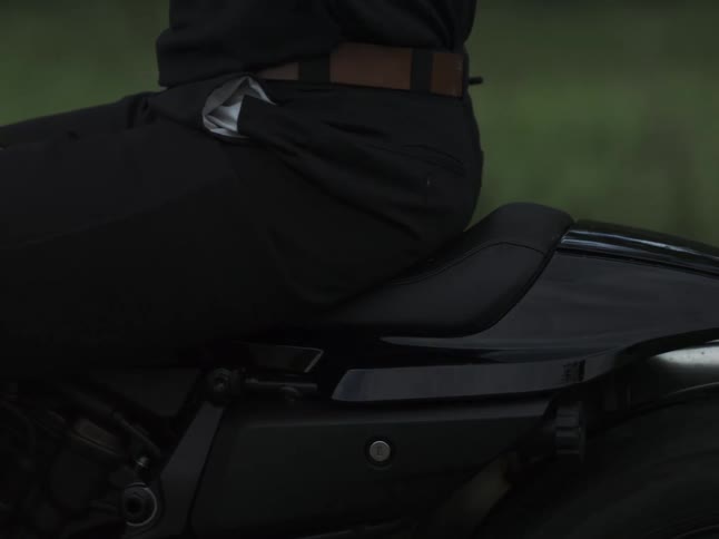 A lateral closeup moves along a black motorcycle from tank to tail as a rider sits poised on the seat belt and pocket lining visible against a blurred green backdrop