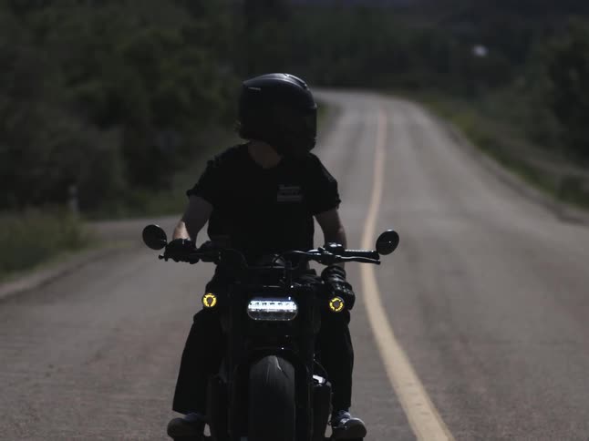 A person rides a motorcycle on a sunny day wearing a helmet steering with one hand while looking toward the side of the road
