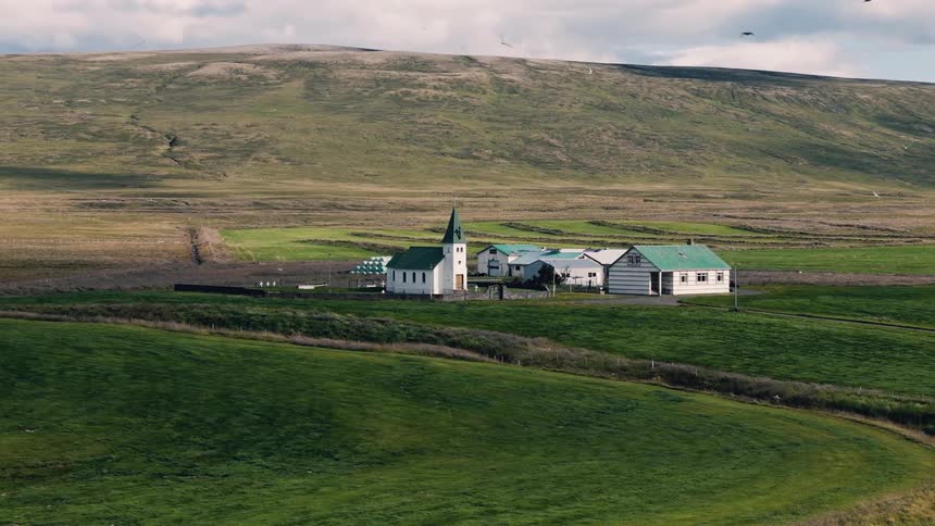 A small rural church complex with green roofs and a pointed steeple sits amid rolling green pastures beneath a broad barren hillside