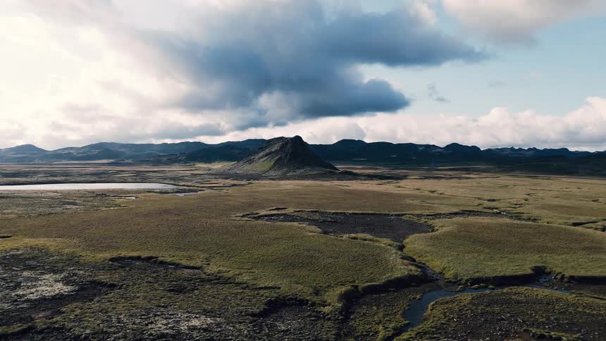 An aerial wide view shows a winding stream flowing through dark green mossy terrain with a dramatic coneshaped rocky mountain rising in the distance under a cloudy dramatic sky