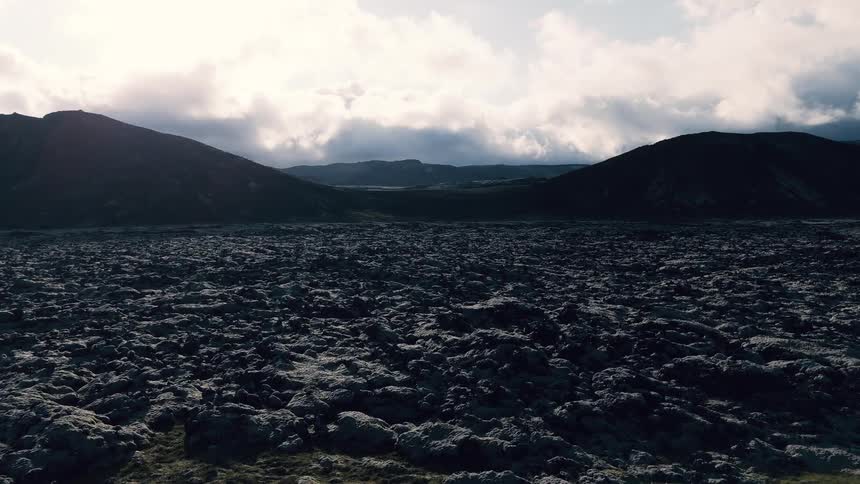 An aerial view moves slowly over a vast dark rocky field of volcanic moss towards two silhouetted hills under an overcast sky with bright patches of light