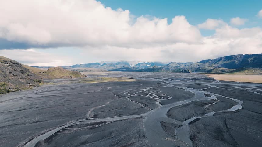 Aerial view of braided glacial channels winding across a vast black sand outwash plain beneath distant mountains and towering clouds
