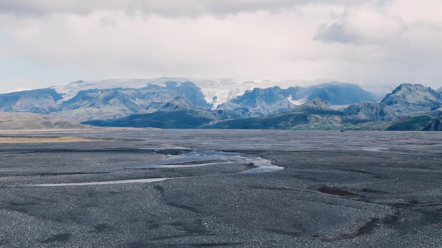 Braided meltwater channels snake across a vast blacksand plain toward jagged glaciercapped mountains under heavy cloud