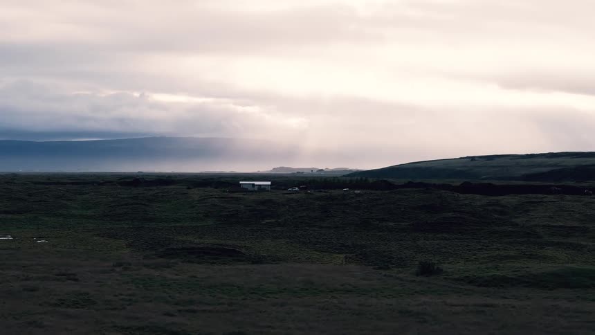 An establishing wide view shows a small white house in the distance nestled in a vast dark green mossy landscape under a dramatic overcast bright sky with rain falling in the background