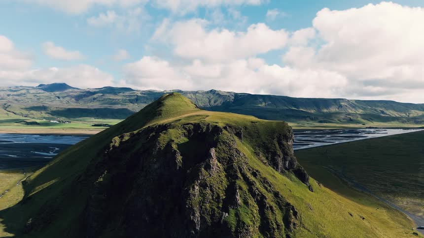 A prominent green conical hill amidst a vast icelandic landscape of flat plains winding rivers and distant mountains under a blue sky with scattered clouds