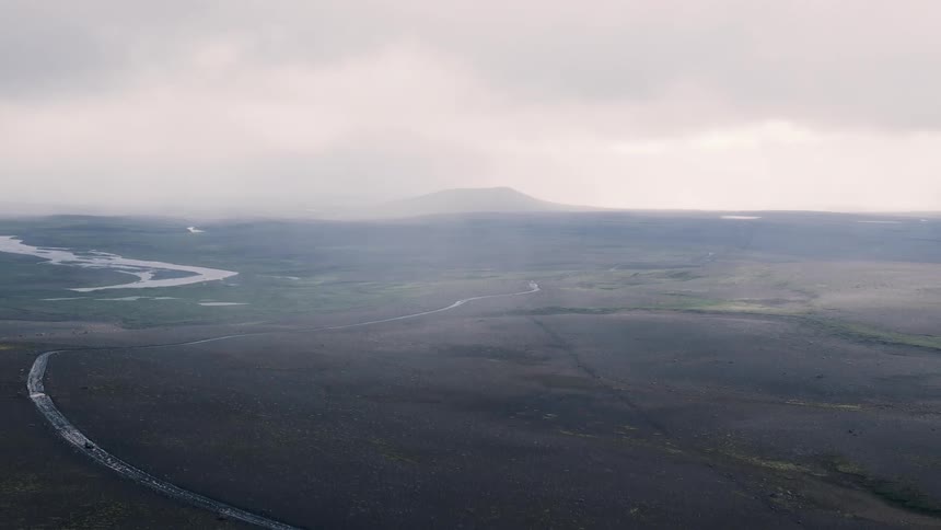 A misty aerial panorama shows a lone road with a vehicle snaking across dark lava plains toward a low hazy mountain with braided rivers glinting under heavy cloud