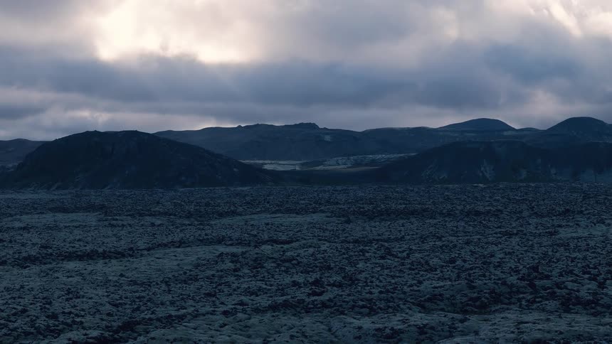 An aerial view moves slowly over a vast dark rocky field of volcanic moss towards a background of silhouetted hills under a dramatic overcast sky with bright patches