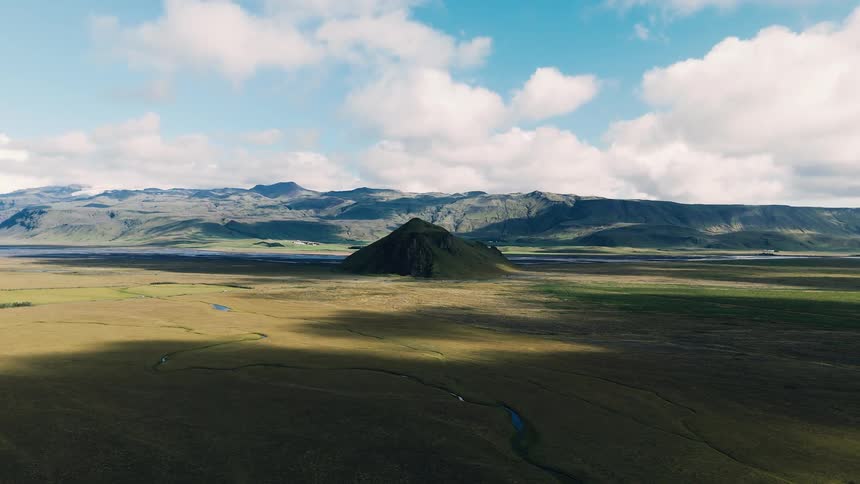 Natural landscape with a plain green area and water streams next to a hill with a mountain range on the background outdoors during day time