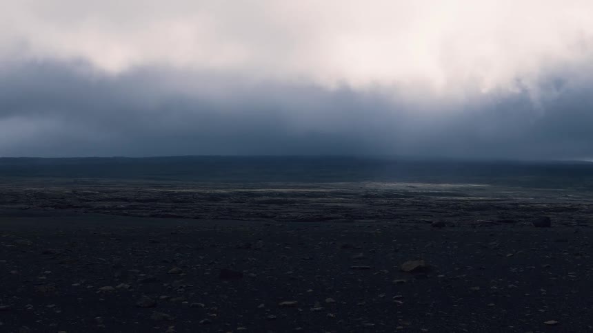 A desolate lava plain of dark rock stretches to a distant ridge under a heavy storm deck with faint sunbreaks along the horizon