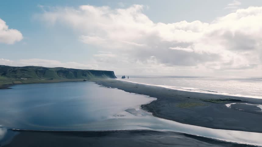 An aerial view shows a calm blue lake and a black sand beach separated from the glittering open ocean by a narrow strip of land with green coastal cliffs and a partly cloudy sky