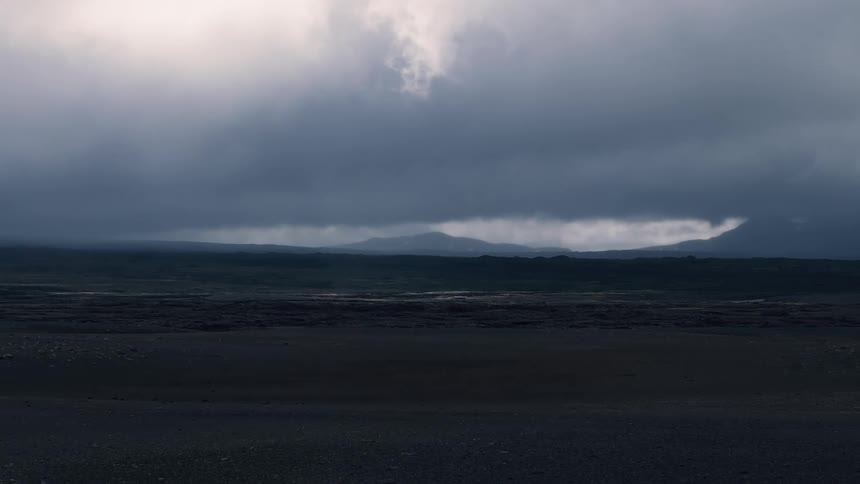 A dramatic wide landscape view shows torrential rain falling from a dark heavy cloud formation onto distant mountains and green plains over a foreground of dark rocky ground