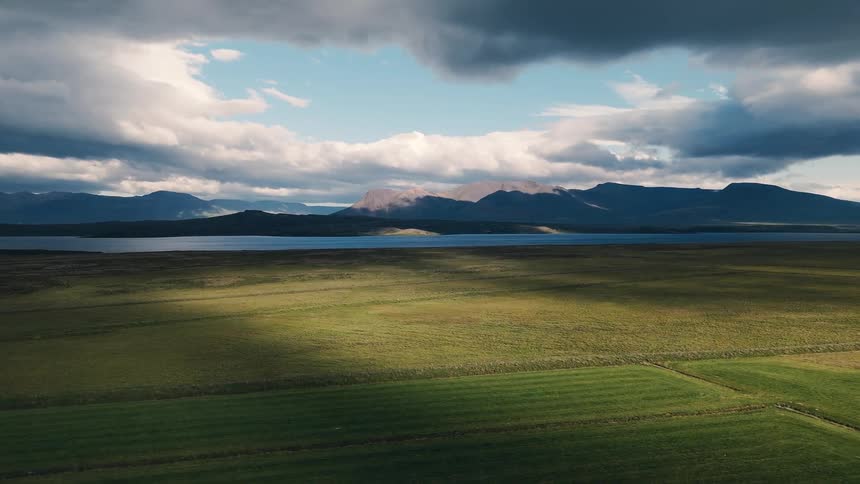 Broad green pastures ripple with drifting cloud shadows beside a long lake backed by blue mountains and towering cumulus
