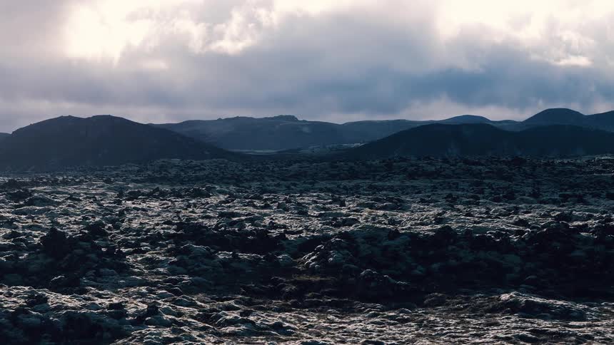 A vast desolate landscape of dark moss covered volcanic rock stretches towards distant hills under a cloudy dramatic sky
