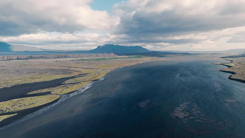 Vast plain with a wide river under dramatic clouds and mountains in the distance