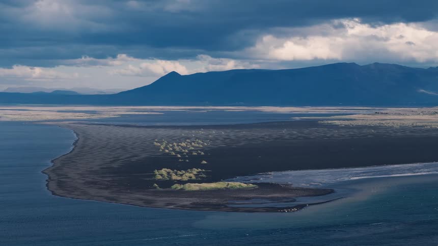 Dark strip of land surrounded by blue waters under a cloudy sky with mountains in the background