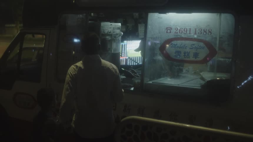 A man and a boy stands at the service window of a parked vehicle illuminated by internal lights displaying phone numbers and text in english and chinese on a dark street with other individuals present