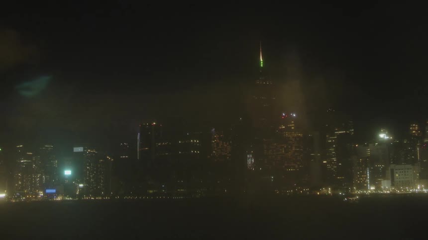 A wide shot shows the illuminated skyline of a modern city at night with a prominent skyscraper in the center and light reflections shimmering on the dark foreground water