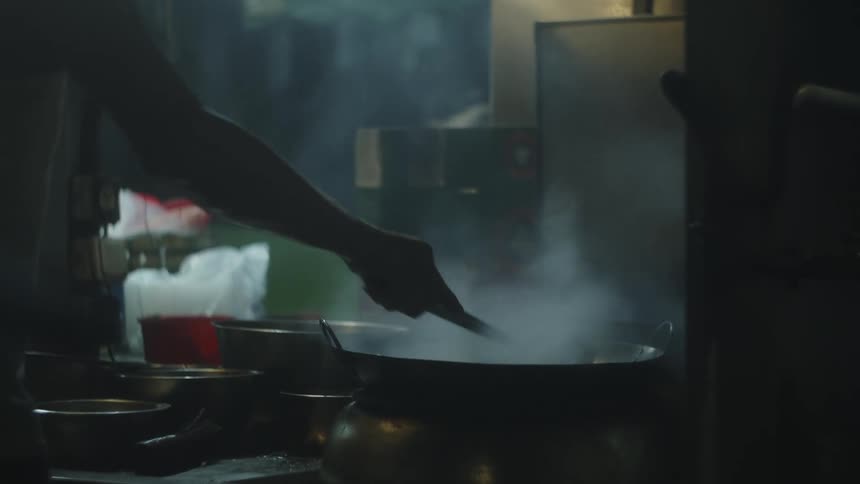 A chef cooks and stirs food in a pan inside the kitchen while another man wearing a hat walks by