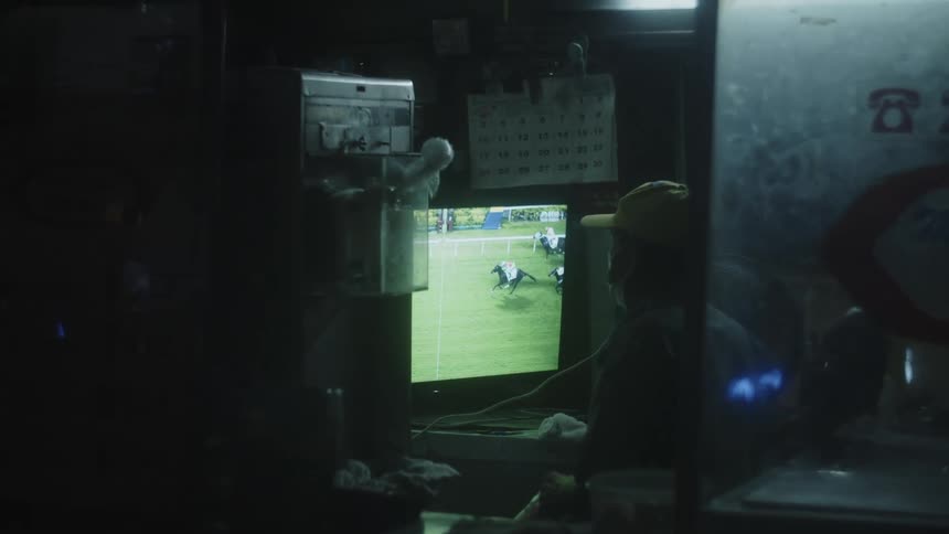 A man wearing a hat watches a horse race on a tv screen inside a cubicle at night