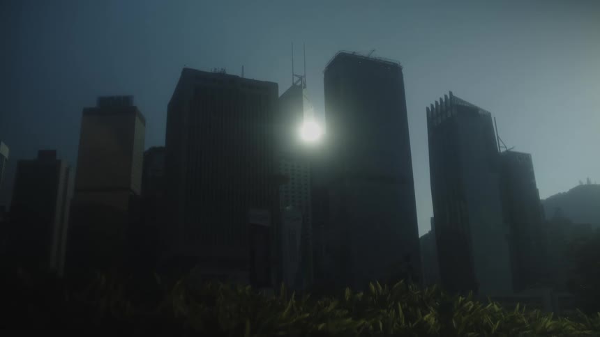 Static lowangle view of multiple towering rectangular structures silhouetted against a bright light source located centrally between two of the buildings with foreground vegetation and a distant hillside visible