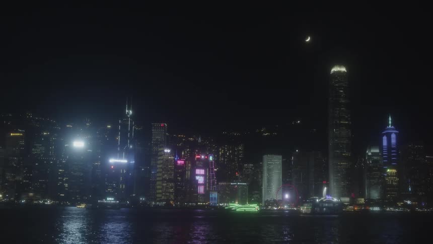 A brightly lit city skyline over water at night with a crescent moon