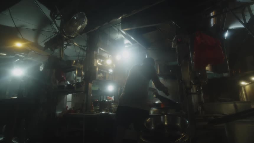 A static lowlight view of multiple figures working within a densely equipped outdoor cooking area or stall with bright light sources and steam obscuring the central action