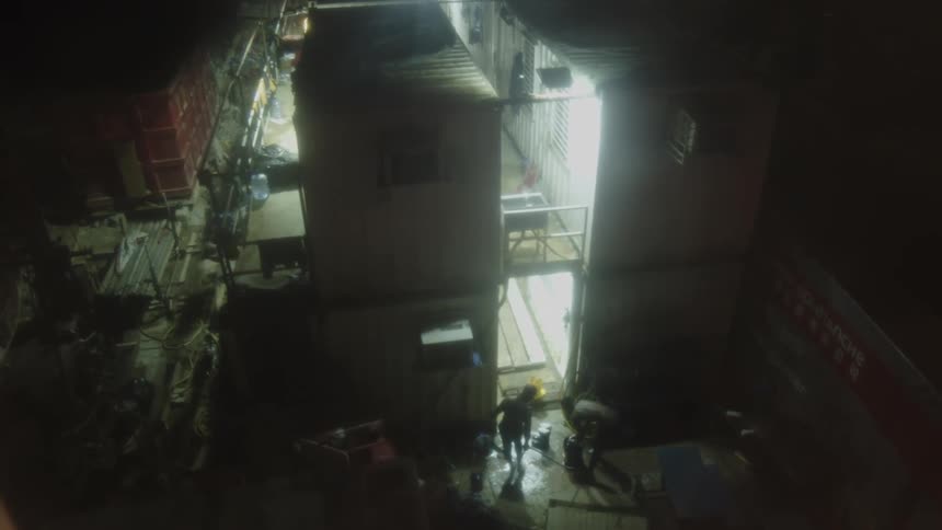 A static highangle view looking down into a confined dark area between temporary structures with two figures moving on a wet floor illuminated by internal lights surrounded by construction materials