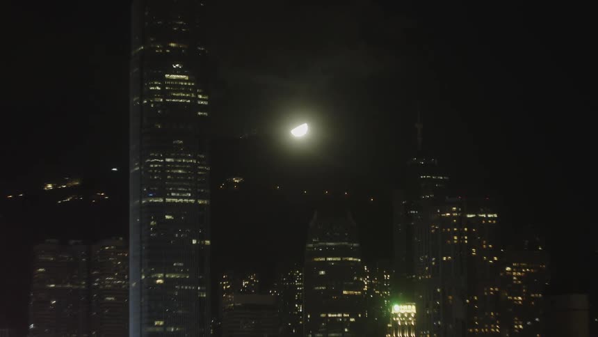 A stationary vertical view of multiple towering structures with many illuminated windows set against a dark night sky where a halfmoon is visible between the buildings