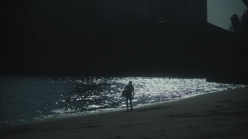 A silhouetted man carrying an object and walking along the edge of a beachm with the water surface next to them reflecting a bright light source