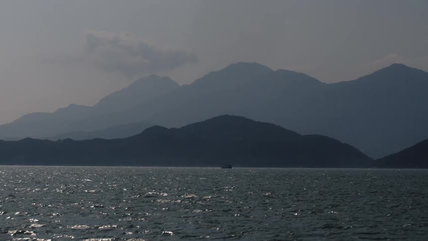 Wide view of a vast expanse of dark agitated water in the foreground with multiple rows of uniformly shaded mountain ridges and a small boat passing by
