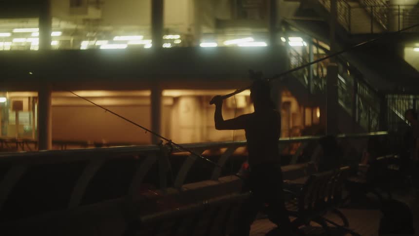 A medium lowlight view of a silhouetted man actively casting a long pole while standing near a railing set against the backdrop of an illuminated multilevel structure at night