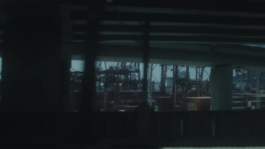 A moving framed view of a large port facility with many vertical cranes and stacked cargo boxes seen through the structural elements of an elevated road or railway line