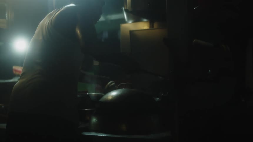 An asian man with their back to the camera stirring contents in a large rounded cooking vessel silhouetted against a bright hazy light source in a cluttered cooking area