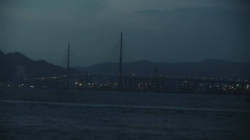 A city bridge glows with lights at night as waves move rapidly in the water below