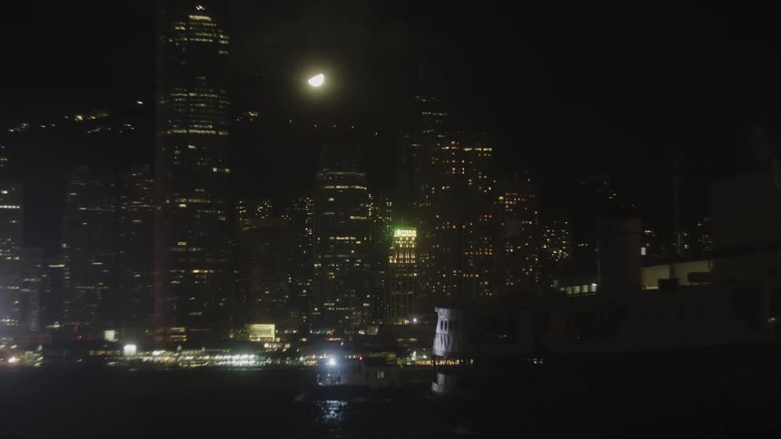 Static wide view of a collection of tall structures with numerous interior lights visible against a dark sky containing the moon while a small vessel moves across the dark body of water in the foreground