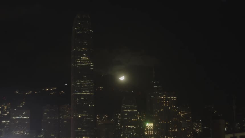 A cityscape with tall skyscrapers lit up and a glowing moon above at night