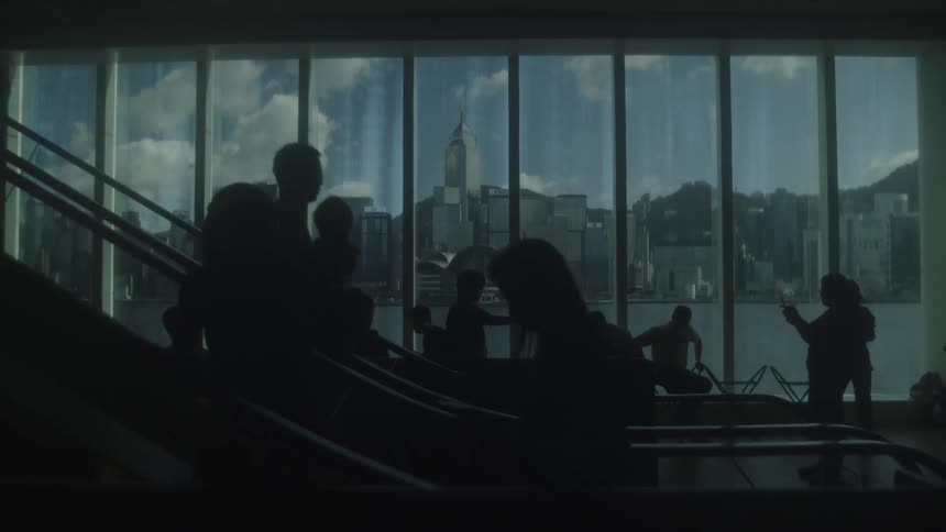 A stationary indoor view of many silhouetted people moving on escalators and standing near a large glass wall that frames a daytime cityscape water and distant hills under a cloudy sky