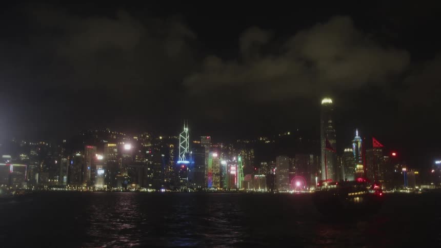 A luxury yacht with illuminated blue lights cruising across a dark body of water against a vibrant city skyline at night