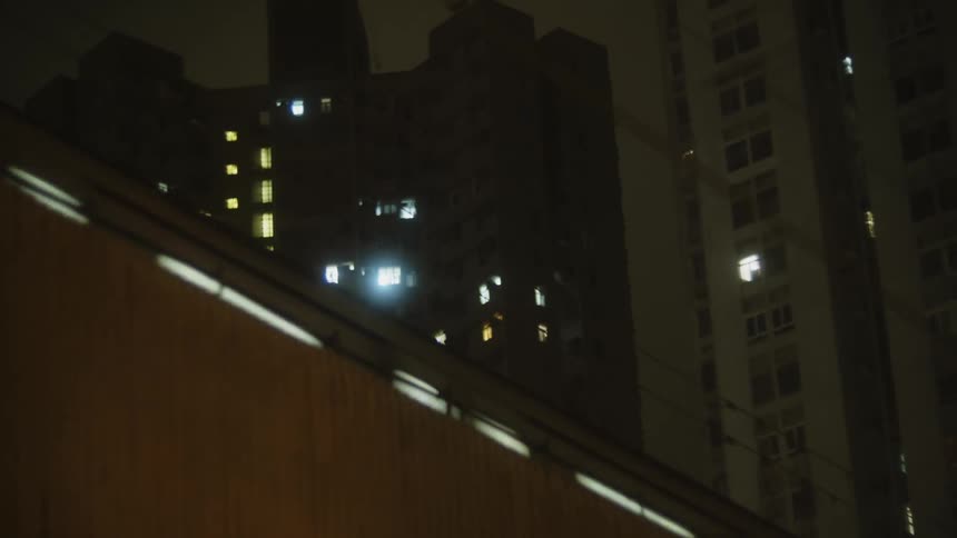An apartment building stands dark against the night sky with scattered windows glowing brightly in a cinematic high angle shot