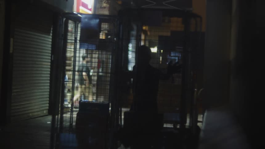 One shadowy figure pushes metal rolling cages down a dark alleyway between closed storefronts illuminated by a distant light source