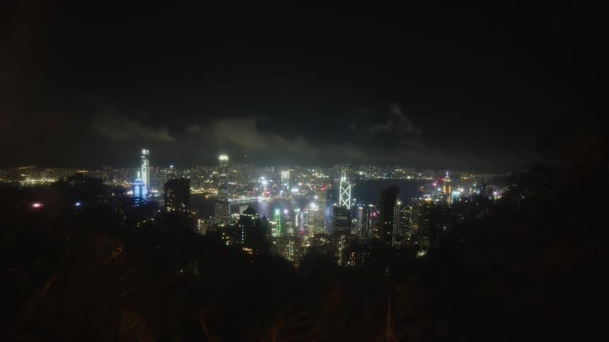 A wide view of a collection of illuminated tall structures and their reflections on the adjacent body of water set beneath a dark sky with clouds framed by dark ground cover in the foreground