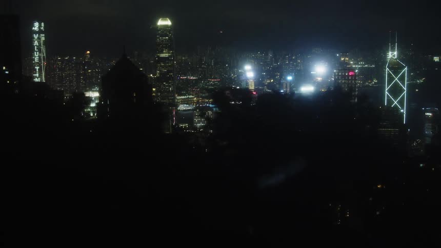 A dynamic highangle view that slowly zooms in on a dense collection of illuminated tall structures at night bordering a dark body of water framed by foliage