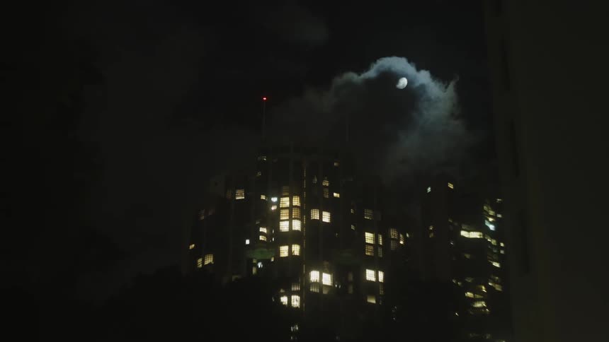 A city skyscraper stands illuminated at night its windows glowing with light as a full moon emerges from behind dark clouds in the sky