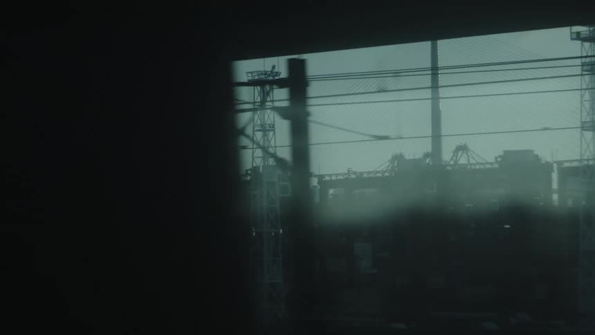 A moving train passes industrial cranes and structures seen through a streaked window